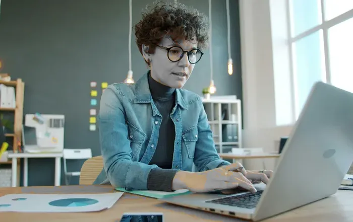 Entrepreneur wearing glasses types on a laptop at a bright modern workspace with papers visible in the background